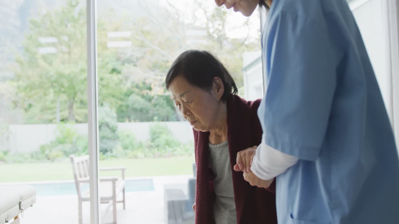 笑顔のアジア人女性医師が、杖を使って高齢の女性患者が歩くのを手伝っている。