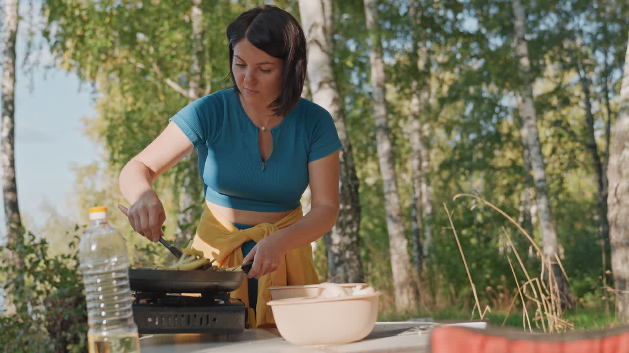 Woman cooking on camping stove, birch trees. Calm lakeside morning with portable gas stove on folding table, blue top and yellow apron, ceramic bowl and water bottle, spatula flipping food in skillet
