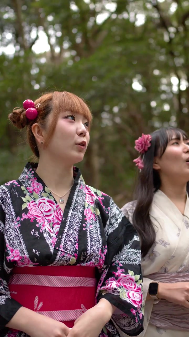 Due donne giapponesi che indossano kimono floreali stanno in una foresta lussureggiante, guardando in alto e sorridendo.