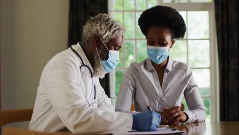 Afroamerikanischer-älterer-Männlicher-Arzt,-Der-Eine-Patientin-Zu-Hause-Besucht-Und-Beide-Gesichtsmasken-Trägt