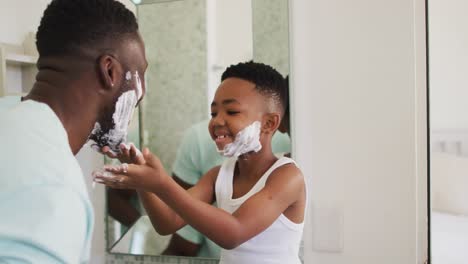 niño afroamericano poniendo crema de afeitar en la cara de su padre y riendo juntos