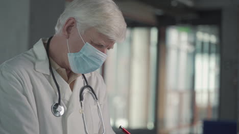 A-Grey-Haired,-Middle-Aged-Doctor-In-A-Facemask-Takes-Notes-In-A-Notebook