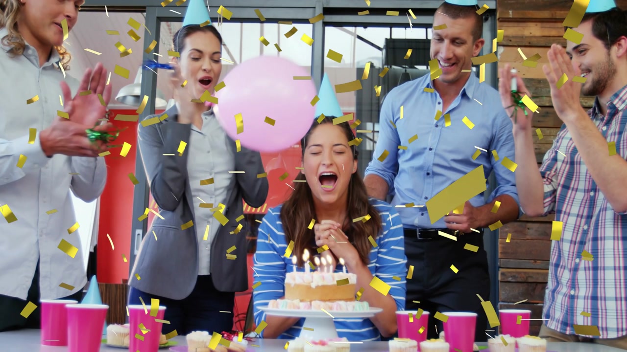 Seated birthday woman blowing cake candles as team popping confetti and balloons for work birthday