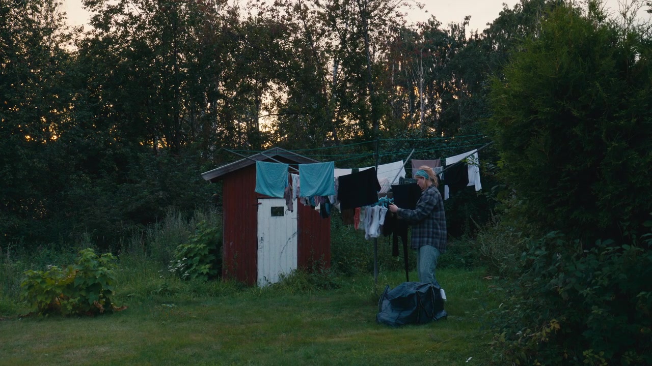 Woman hanging freshly washed clothes on an outdoor clothesline in a quiet country garden at dusk, celebrating slow living, sustainability and simple everyday routines.