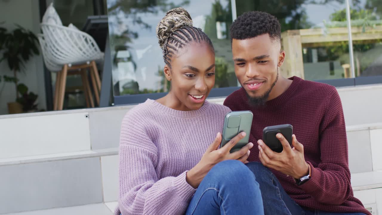 Happy african american couple talking and relaxing with smartphones outside house