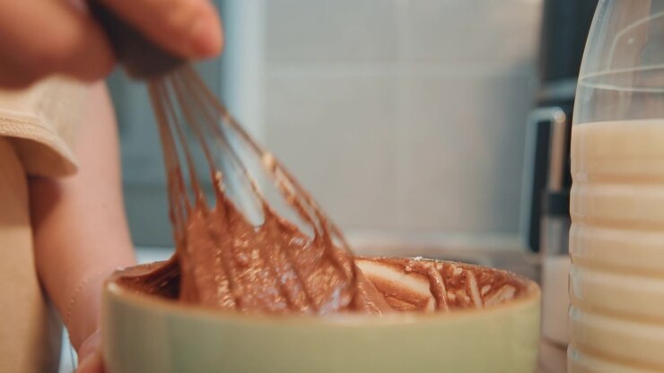 Closeup whisking chocolate batter in bowl, green ceramic bowl and metal whisk, milk jug at edge, warm kitchen tiles, home cook prepping dessert with steady rhythmic strokes, sticky cocoa texture