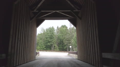 Pushing-slowly-forward-towards-the-road-through-the-dark,-dramatic-tunnel-of-the-public-Lowe's-covered-bridge-in-Maine-with-a-logging-truck-passing-quickly-by