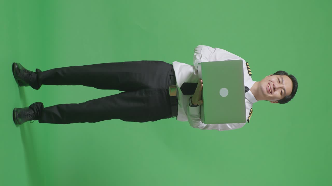 cuerpo lleno de piloto de hombre asiático usando una computadora portátil y sonriendo a la cámara mientras está de pie en el estudio de fondo de la pantalla verde