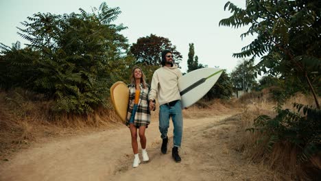 A-guy-in-a-hat-and-sunglasses-in-a-white-sweatshirt-walks-with-his-blonde-girlfriend-in-a-plaid-shirt-holding-hands-and-they-carry-surfboards-along-the-shore-near-the-sea.-Guy-and-girl-Surfers-go-down-the-slope-to-ride-and-swim-in-the-sea