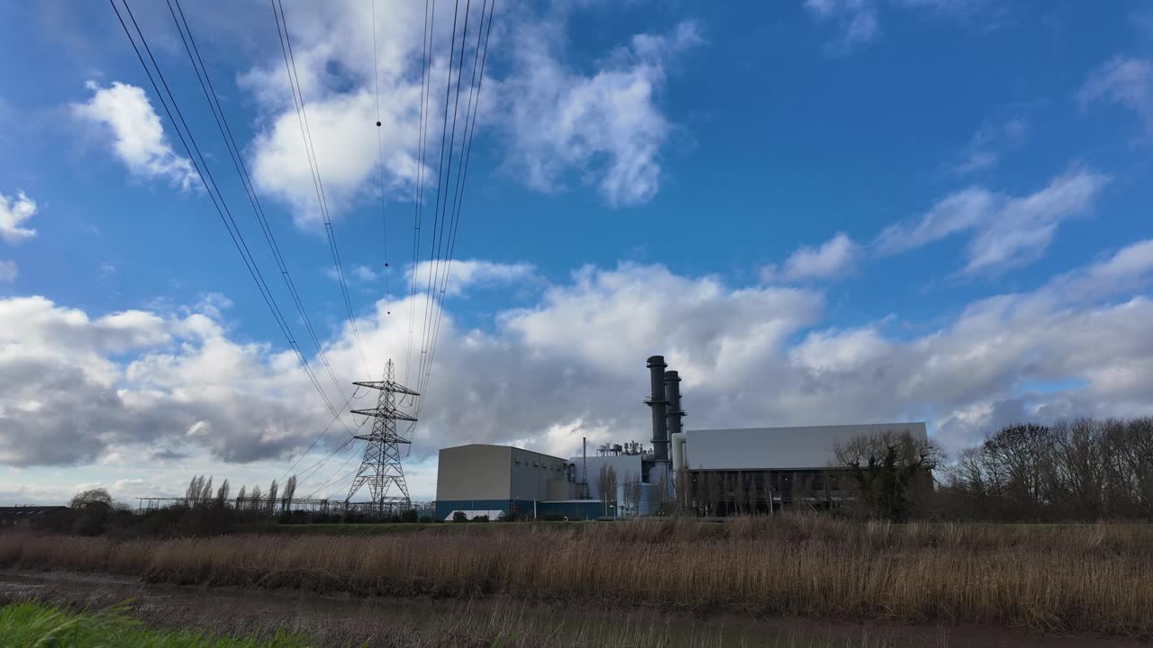 Industrial plant with smokestack and power lines in rural field under cloudy sky
