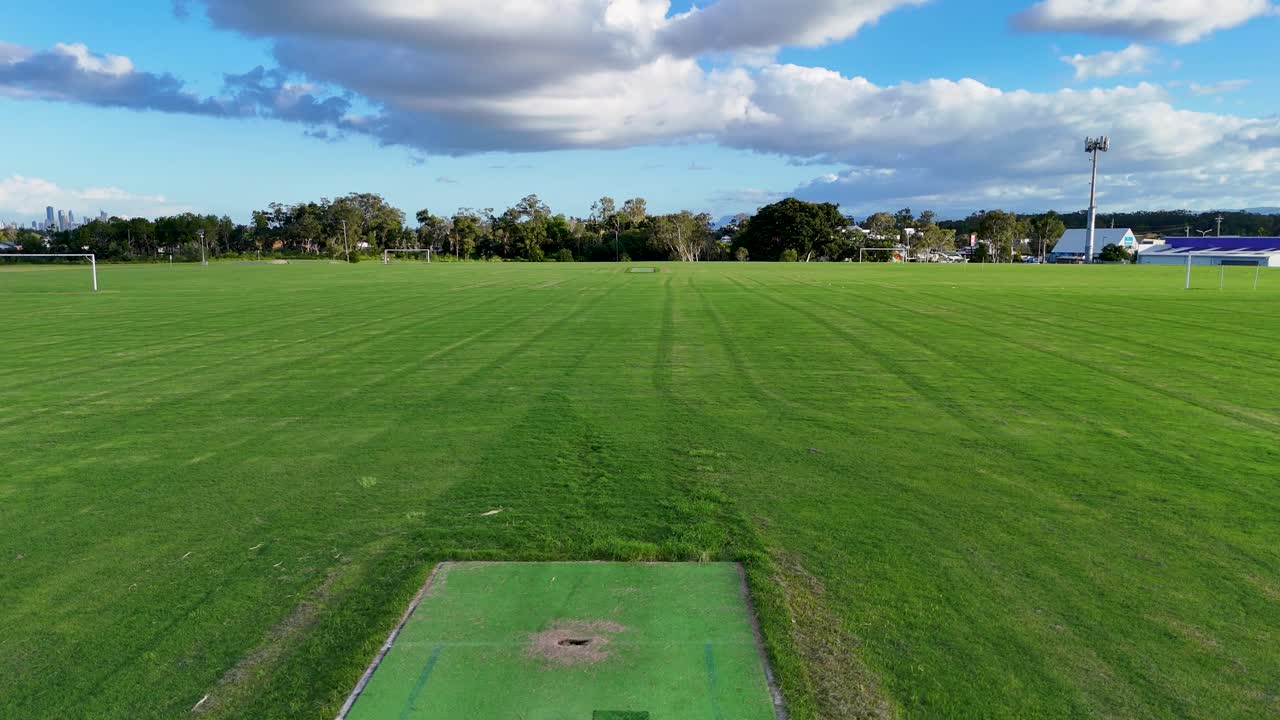 Aerial view of a sports field with a cricket pitch, captured in bright daylight, showcasing expansive green grass and clear skies