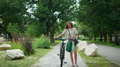 young adult strolling with bicycle, holding handle with green and mint bags attached, adjusting black handbag, surrounded by lush greenery and rocks along park pathway