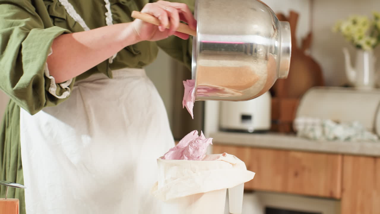 Chef scrapes fluffy pink dough from large mixing bowl into piping bag using wooden spatula, preparing for baking in cozy kitchen setting with warm lighting and rustic decor