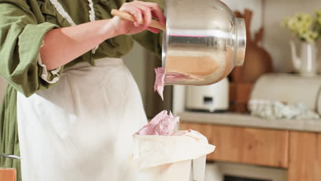 chef scrapes fluffy pink dough from large mixing bowl into piping bag using wooden spatula, preparing for baking in cozy kitchen setting with warm lighting and rustic decor