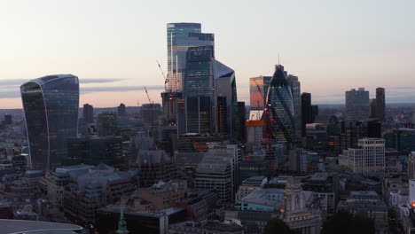 Rising-footage-of-group-of-modern-futuristic-office-downtown-buildings.-Gherkin,-Scalpel,-Walkie-Talkie,-Leadenhall-and-other-iconic-skyscrapers-against-bright-sky-after-sunset.-London,-UK