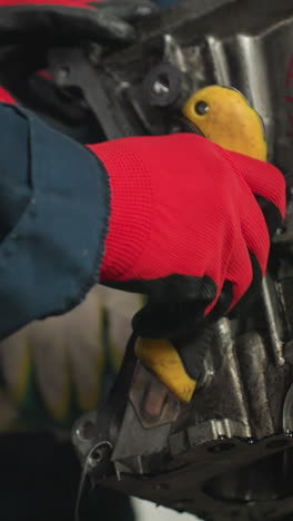 primer plano de un mecánico en uniforme azul usando un raspador amarillo en un motor de coche, otro mecánico con guantes rojos ayuda, en un taller automotriz ocupado