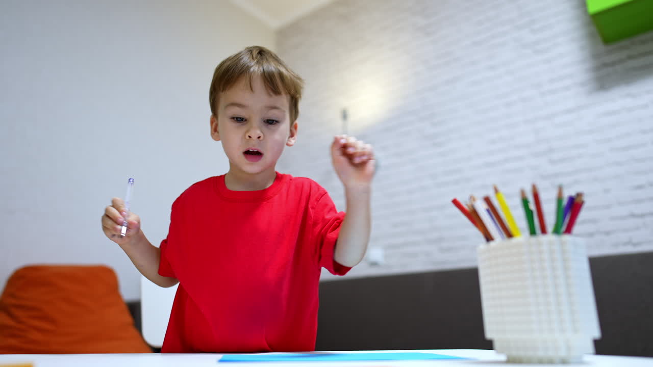 Joyful toddler boy in red t-shirt holds a pen. Lovely baby draws cheerfully at home. Low angle view.