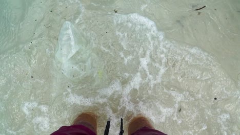 vista desde arriba en primera persona de las piernas y las olas del mar contaminadas bañando los pies descalzos.