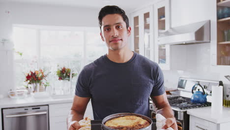 Un-Hombre-Hispano-Milenario-Decepcionado-Parado-En-La-Cocina-Presentando-El-Pastel-Que-Ha-Horneado-A-La-Cámara