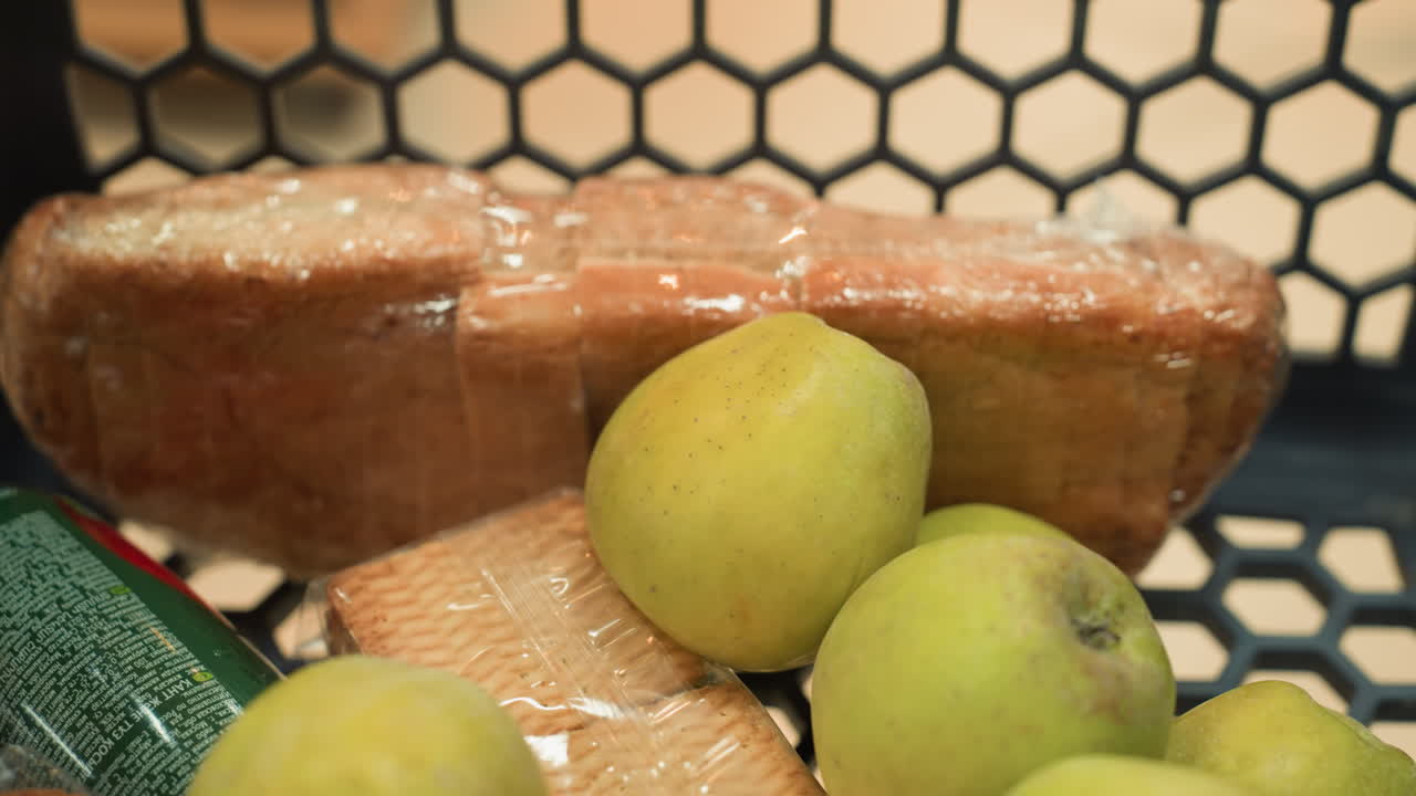 vista de cerca de una mano colocando plátanos en un carrito de compras que ya contiene un paquete de galletas, un pequeño jugo, un pan y varias manzanas