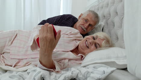 Senior-grandparents-couple-lying-in-bed.-Woman-getting-nervous-about-man-spying-into-mobile-phone