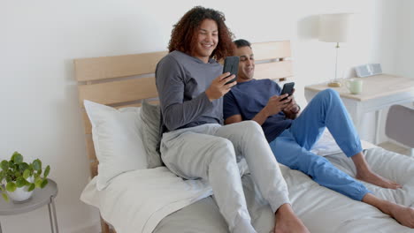 happy diverse gay male couple sitting on bed using smartphones in the morning, slow motion