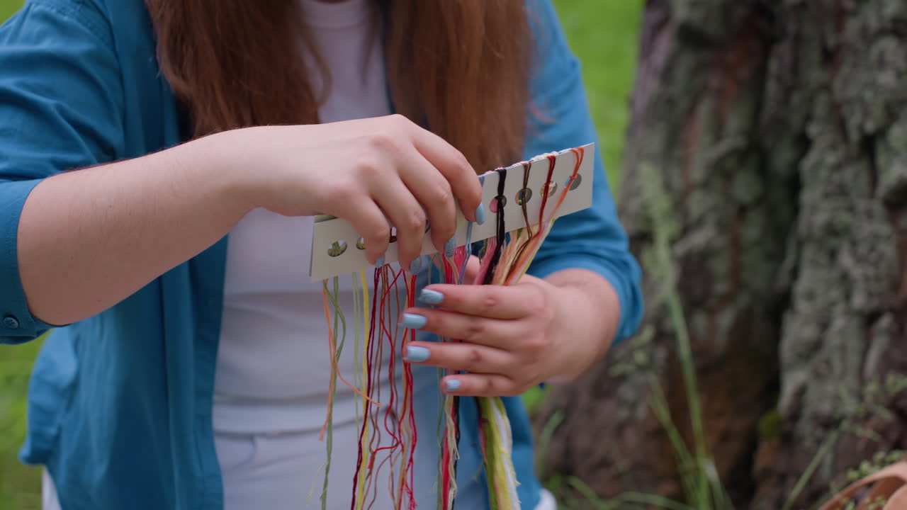 Young girl drops colorful embroidery threads on blanket with water bottle and banana nearby, enjoying peaceful crafting moment surrounded by fresh green grass and natural light
