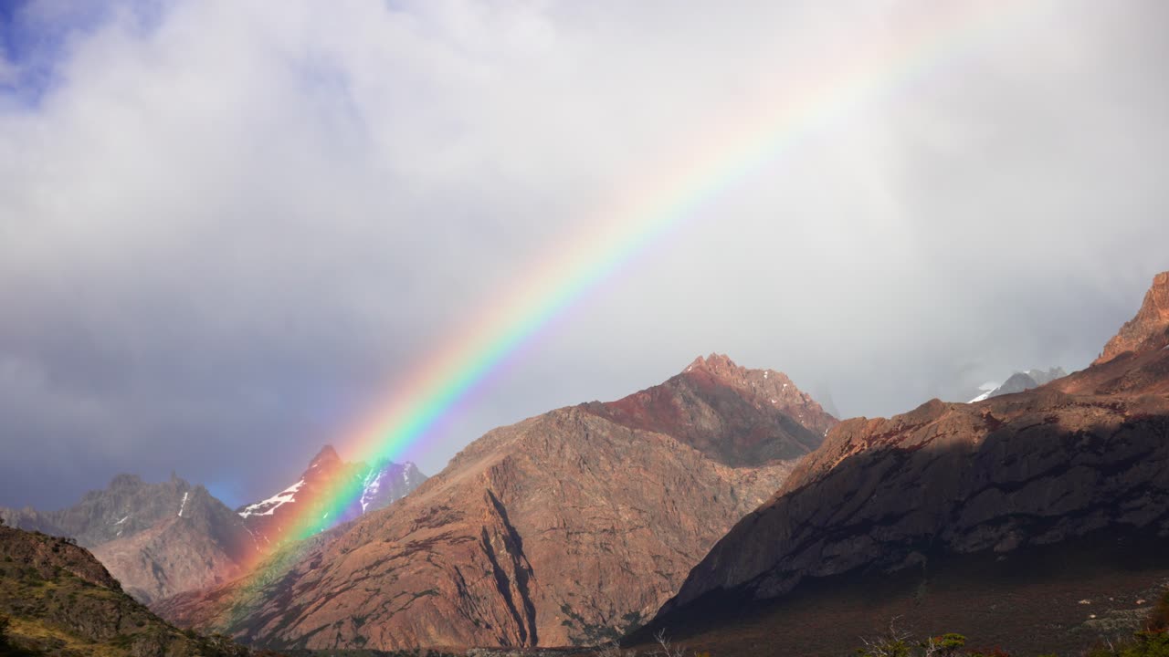 Timelapse view of a vibrant rainbow over the Andes Mountains in Patagonia with clouds casting moving shadows across the peaks