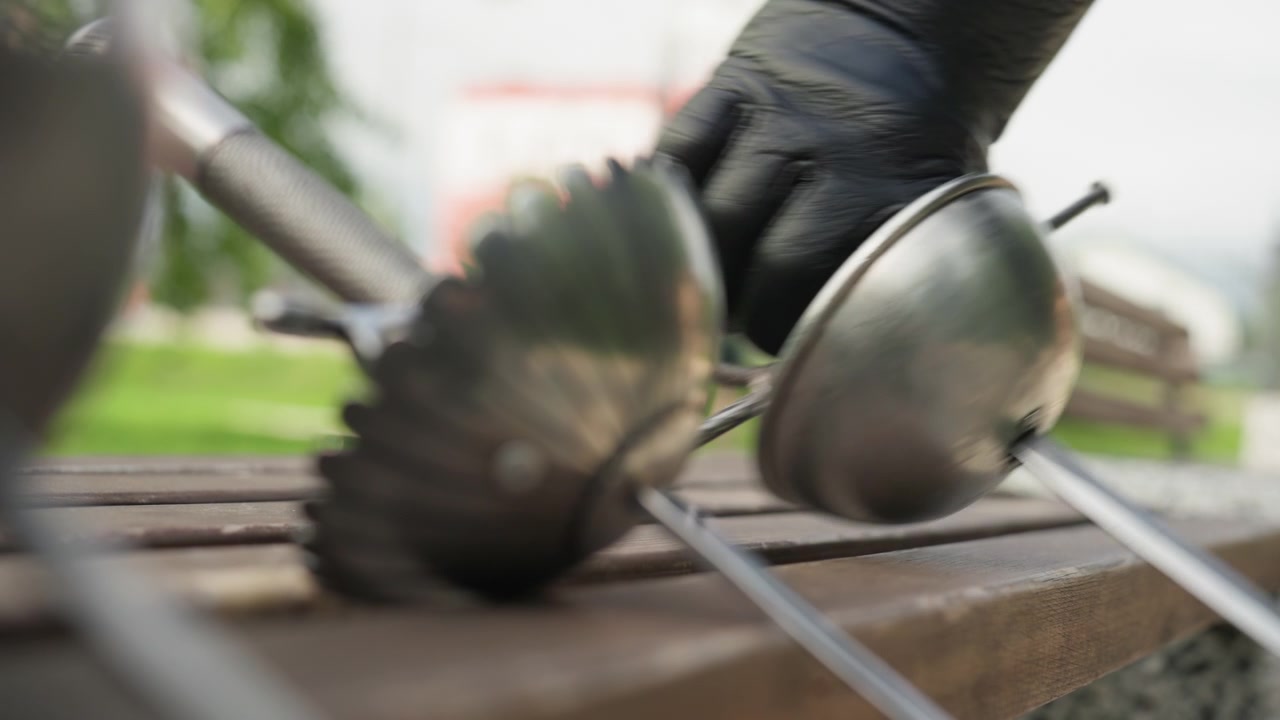 Close up of two crossed fencing swords on wooden bench with soft focus background. gloved hand briefly interacts by adjusting one sword emphasizing metallic texture and historical design