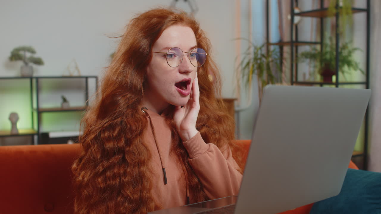 Surprised caucasian redhead woman with windswept hair using laptop sitting on couch at home