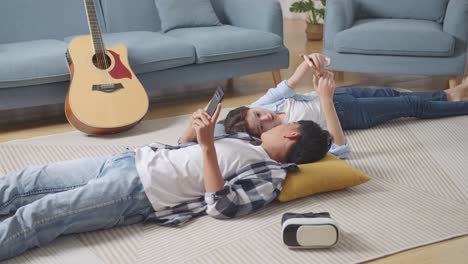 asian teen couple playing smartphone while lying on carpet on the floor at home. phone addicted couple speaking, laughing, and enjoying time together