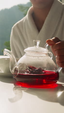 man puts teapot with tea on table. young people enjoy morning tea-time sitting together in kitchen by window. process of morning tea ceremony