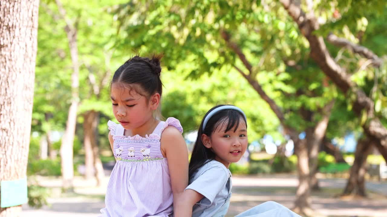 สองพี่น้องชาวเอเชียวัยเยาว์นั่งหันหลังชนกันกลางแจ้งในสวนสาธารณะสีเขียวสดใสที่มีแดดจ้า