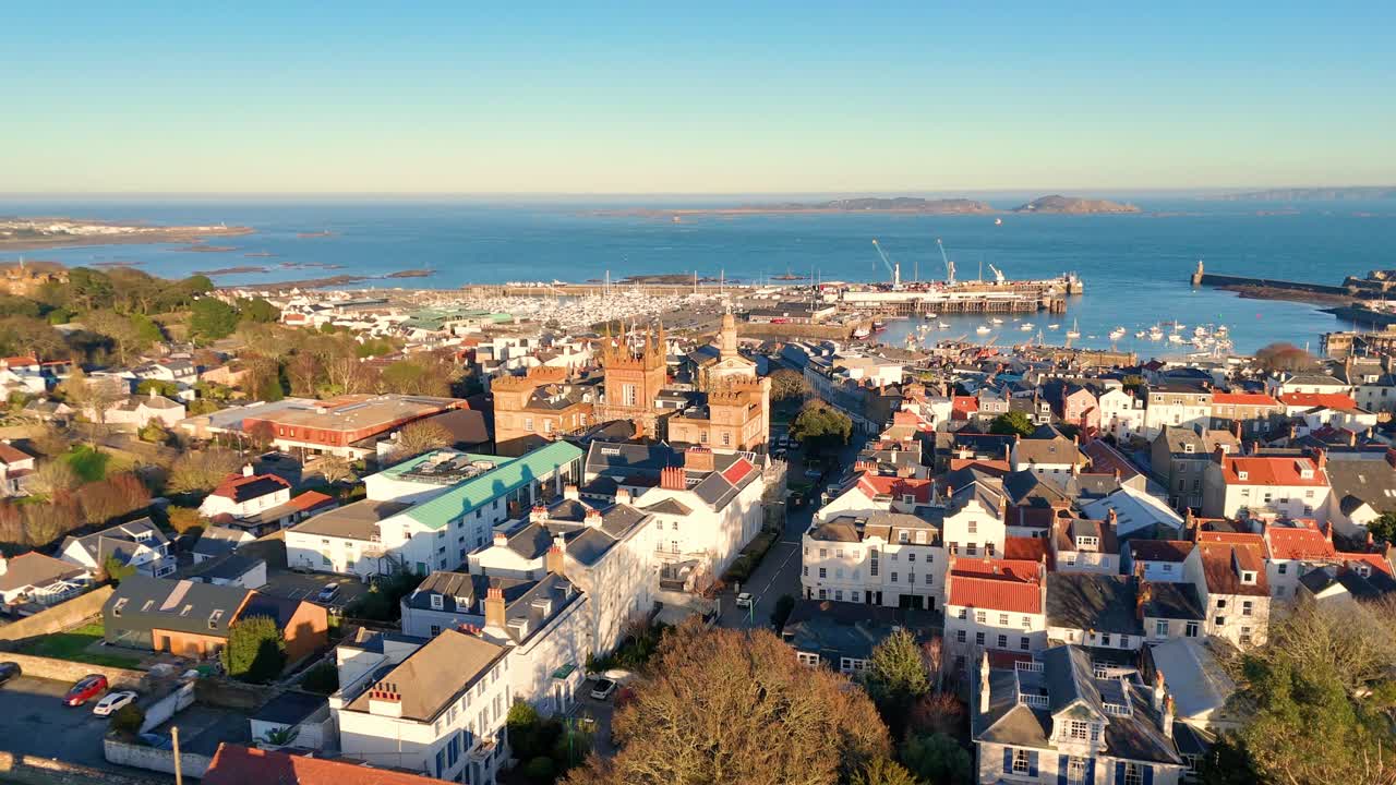 High backwards drone footage of the roof tops of St Peter Port Guernsey in the golden hour with extensive views across town and the harbour to Herm and Jethou