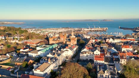 high backwards drone footage of the roof tops of st peter port guernsey in the golden hour with extensive views across town and the harbour to herm and jethou