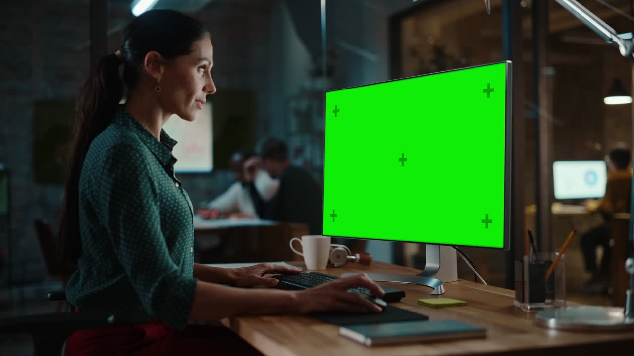 Middle Aged Multiethnic Specialist Working on Desktop Computer with Green Screen Mock Up Display in Busy Creative Office. Beautiful Diverse Female Manager in Green Polka Dot Blouse.