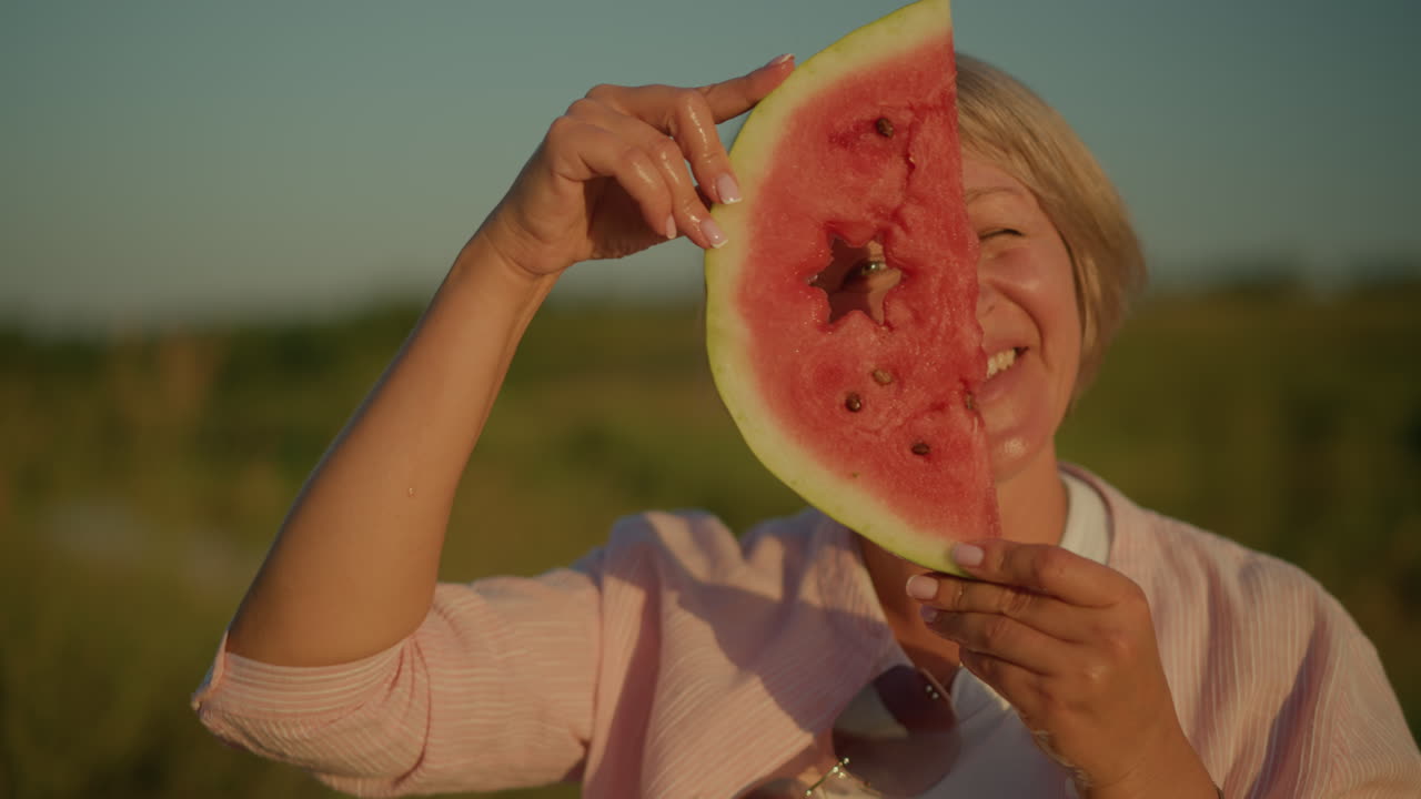 mujer sosteniendo una rebanada de sandía con un recorte en forma de estrella mientras sonríe juguetón, mirando a través del recorte bajo la luz del sol brillante, el fondo presenta un paisaje verde borroso