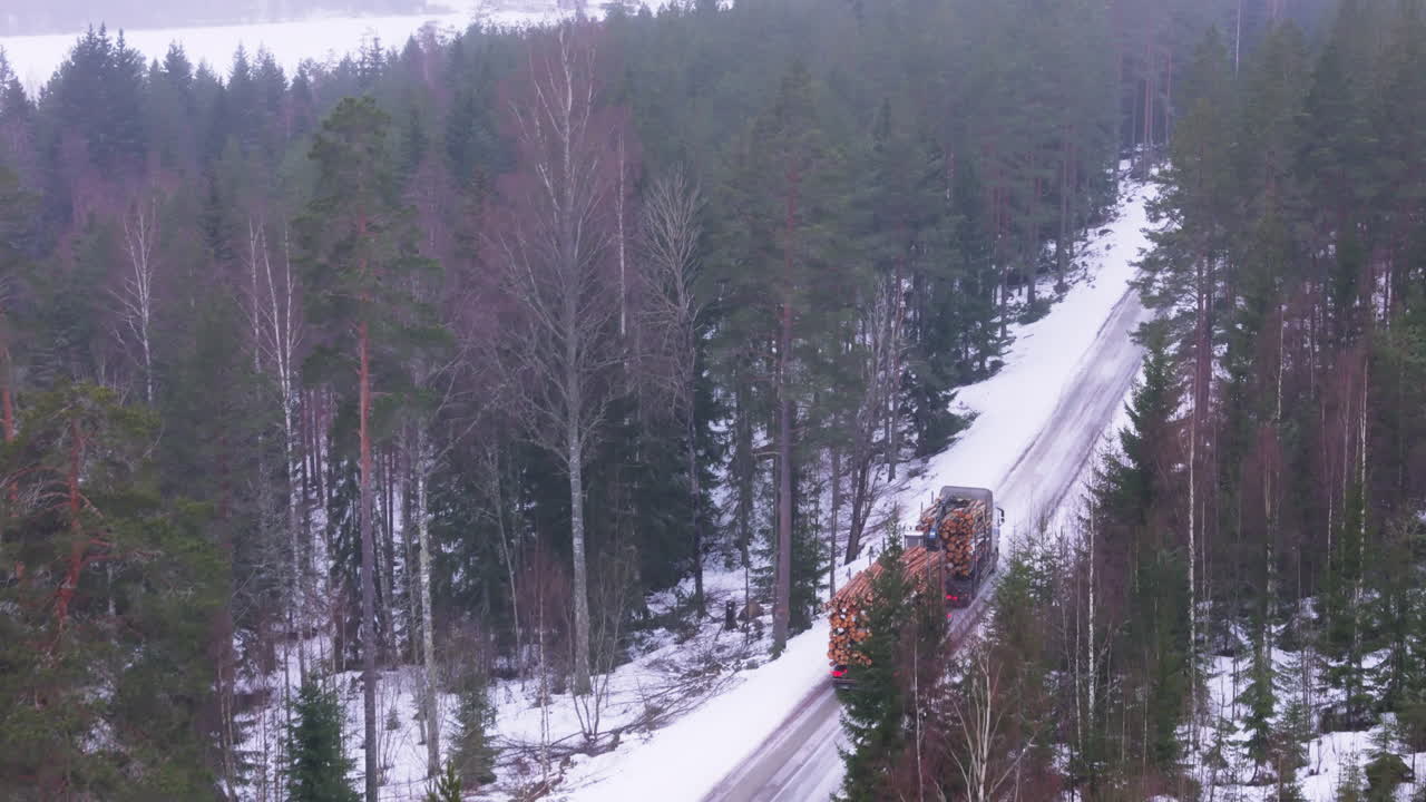 Truck transports harvested timber logs on snowy forest road, telephoto aerial