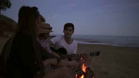 Group-Of-Young-And-Cheerful-People-Sitting-By-The-Fire-On-The-Beach-In-The-Evening,-Grilling-Sausages-And-Playing-Guitar-1