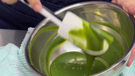 primer plano de un pastelero profesional mezclando y preparando a mano un brillante glaseado de espejo de chocolate blanco matcha con una espátula en una panadería de cocina comercial