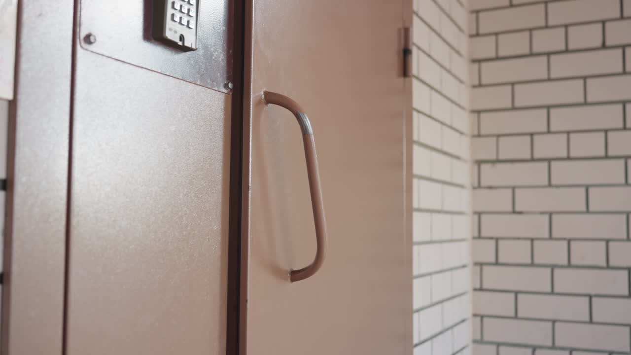 Escena de entrada de seguridad que muestra una puerta de metal pesada con teclado y una manija grande que se cierra de golpe mientras una empresaria pasa por delante, las bisagras hacen ruido, las paredes del pasillo y los ladrillos son visibles, control que sugiere la entrada