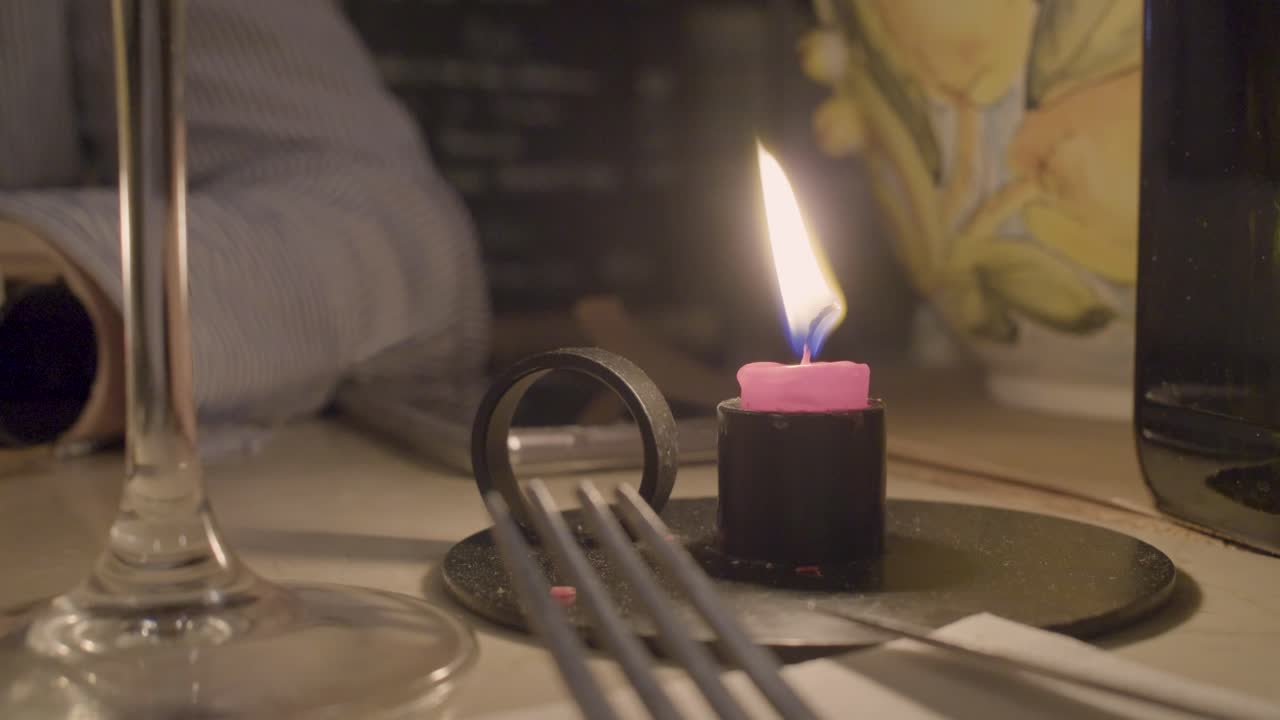 A close-up shot of a pink candle burning brightly next to some utensils and a glass