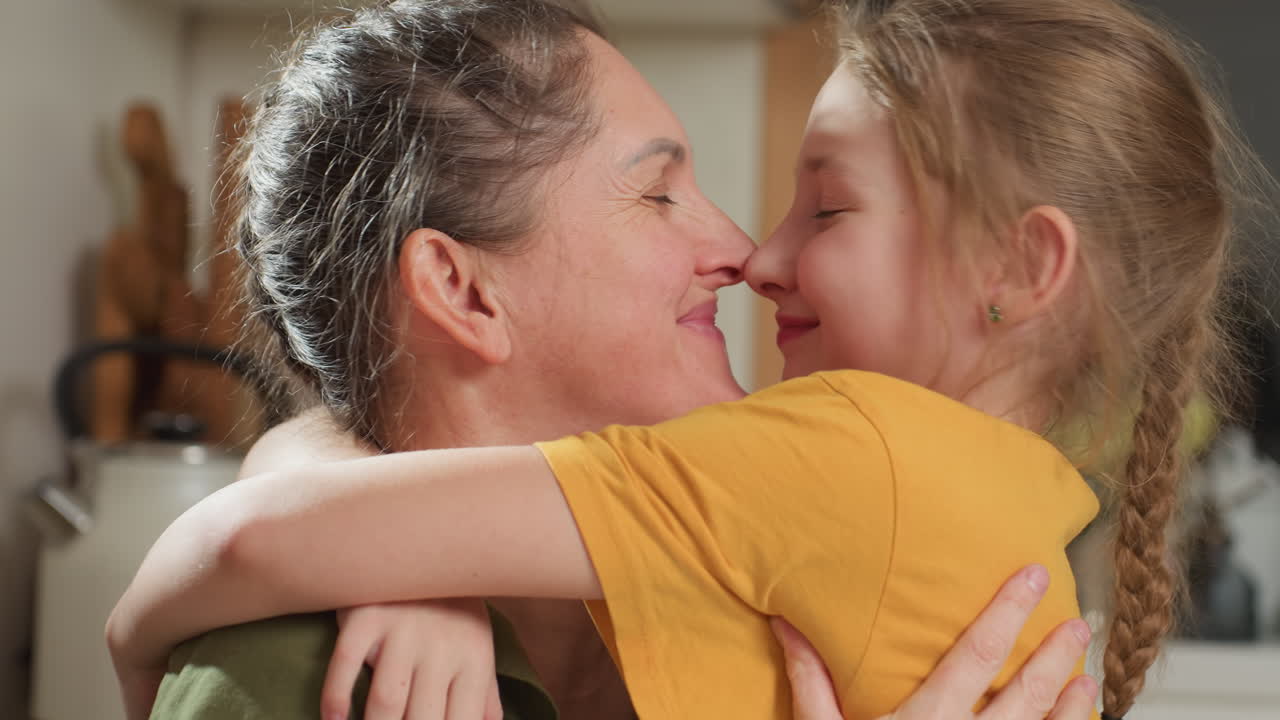 Joyful mother and daughter affectionately embrace in warm kitchen setting, smiling with eyes closed while playfully rubbing noses, expressing deep love, trust, and bonding