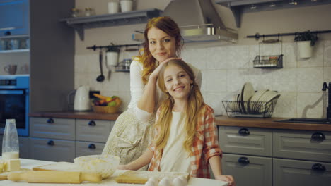 Portrait-shot-of-the-young-mother-and-her-cute-daughter-standing-closely,-looking-at-each-other-and-thanto-the-camera-in-the-kitchen-at-the-table-while-baking.-Indoors