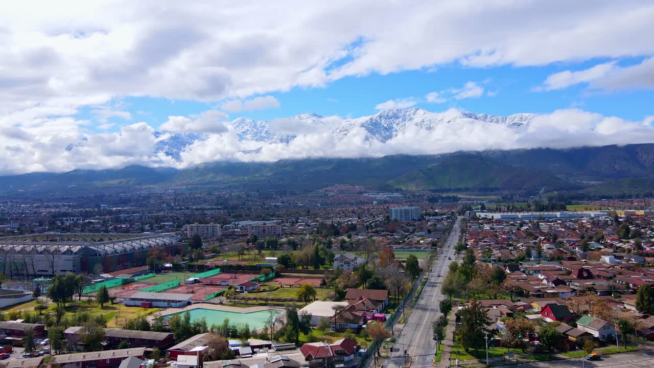 Volo aereo in avanti sopra un quartiere residenziale e impianti sportivi verso le maestose Ande innevate, parzialmente coperte dalle nuvole sotto un cielo blu. Santiago, Cile.