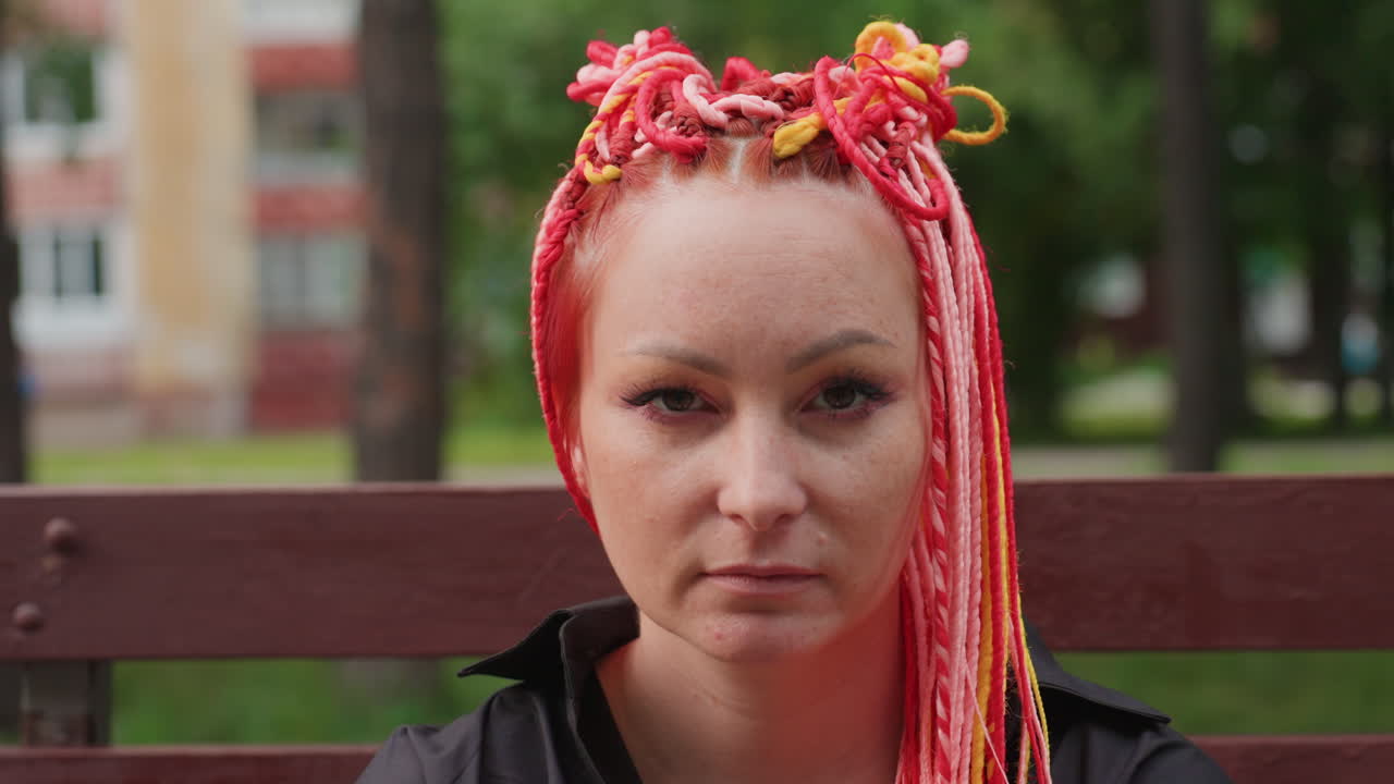 primer plano de músico callejero caucásico sentado en un banco, rastas coloridas enmarcando un rostro solemne, mirada directa a la cámara, sutiles pecas y maquillaje, parque urbano de fondo, ambiente introspectivo y silencioso