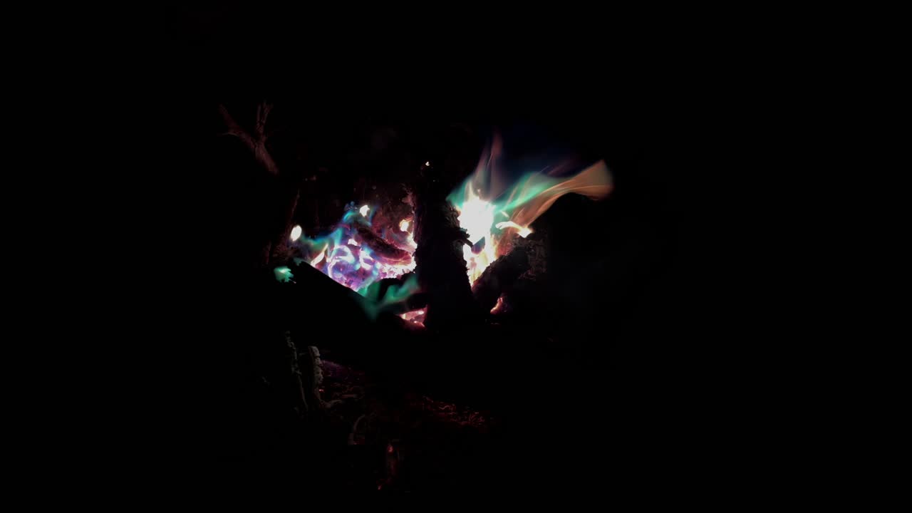 Atmospheric close-up footage of a campfire with flickering flames and glowing embers in deep darkness at night, creating warm light, heat, and cozy outdoor mood