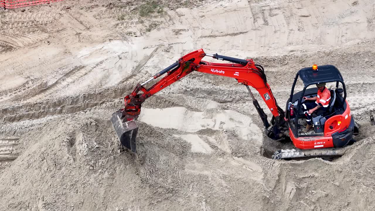 En rød minigraver graver og stabler sand på en solrig australsk byggeplads ved vandet