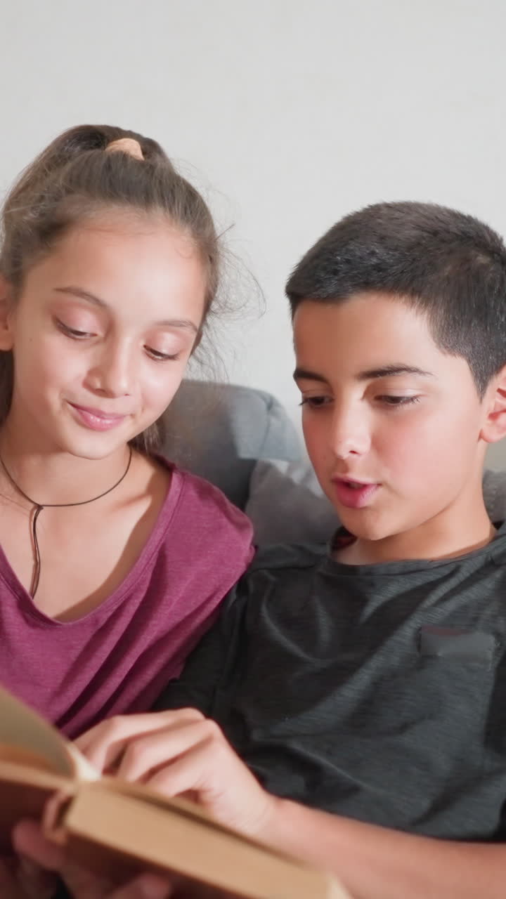 Momento di apprendimento interattivo tra fratelli che leggono un libro insieme sul divano con sorrisi e interesse. Il ragazzo legge ad alta voce mentre la ragazza ascolta tenendo in mano una penna, entrambi coinvolti