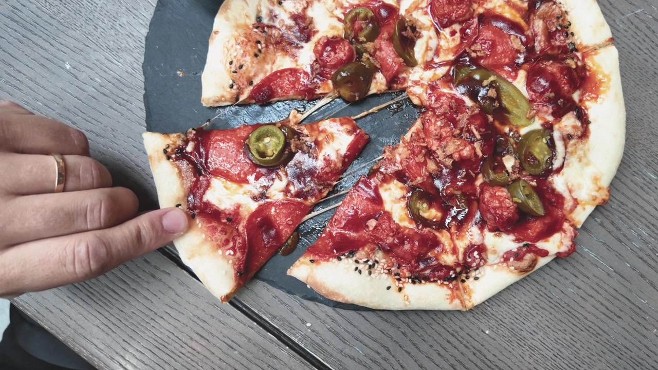 Una persona afferra una fetta calda e filante di pizza al salame piccante e jalapeño da un tavolo di legno rustico in un primo piano. Il formaggio fuso si allunga mentre la fetta di pizza viene sollevata, mostrando deliziosi dettagli